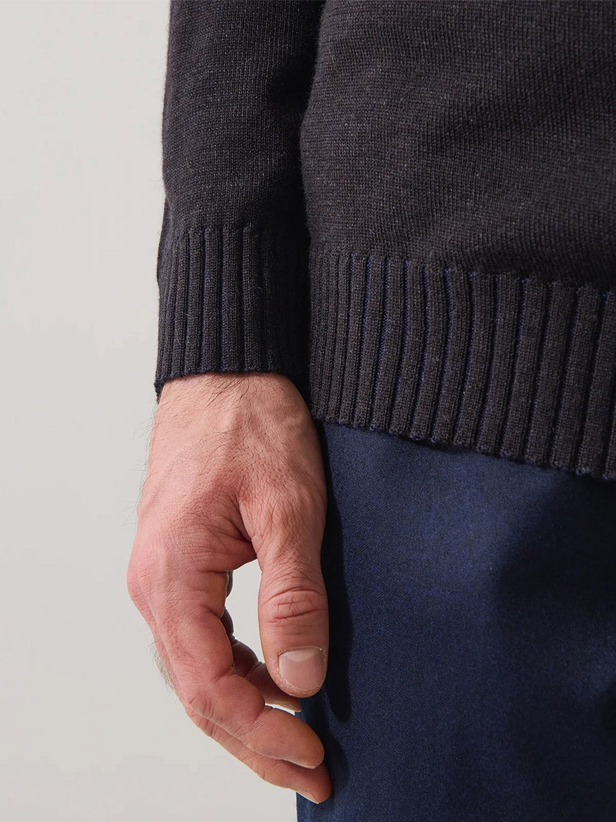A close-up of a person’s left hand resting beside their dark navy pants, wearing the Patrick Assaraf Merino Plaited Quarter Zip in Brownie—a dark knit sweater with a ribbed cuff that offers lightweight warmth and a refined silhouette.
