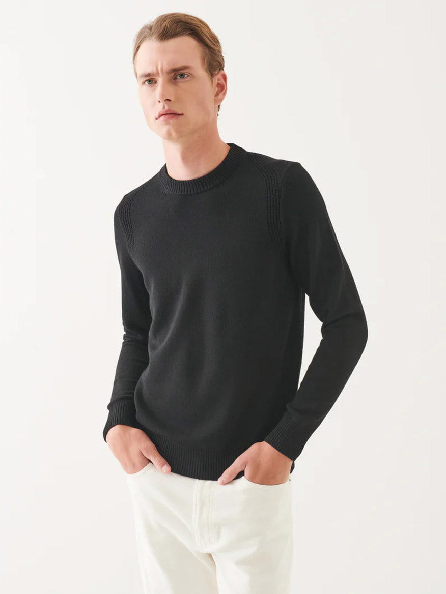 A person wearing the Patrick Assaraf Merino Crewneck in Black paired with white pants stands against a plain white background, hands in pockets.