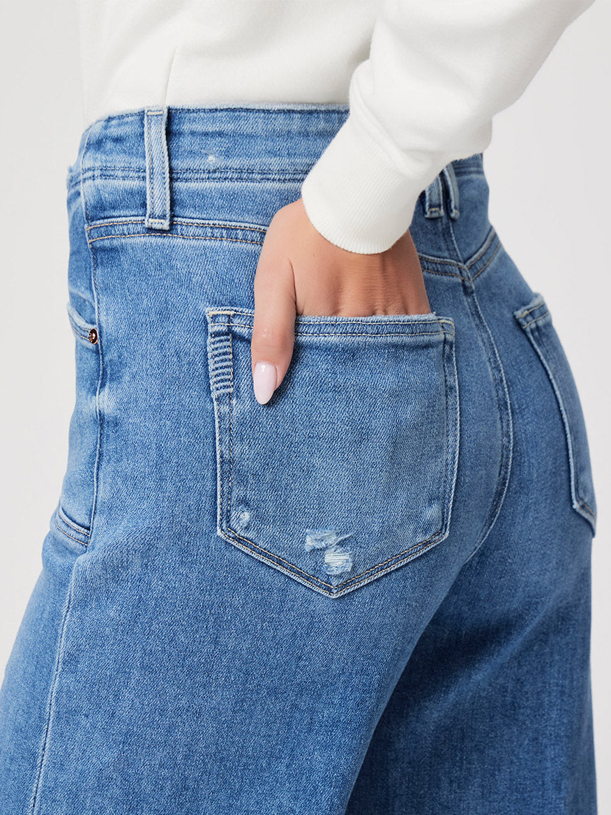 Close-up of a person wearing Paige Anessa Wide Leg in Sunnie Distressed, with a hand partially in the back pocket. The light blue jeans feature a raw hem and are paired with a white long-sleeve top.