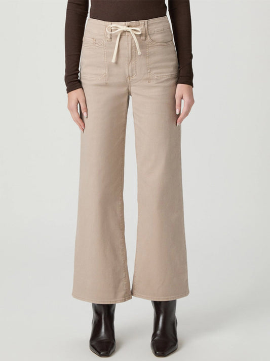 Wearing the Paige Ari Wide Leg Pant in Vintage Utopia, paired with a brown long-sleeve top and dark ankle boots, a person stands against a plain background.