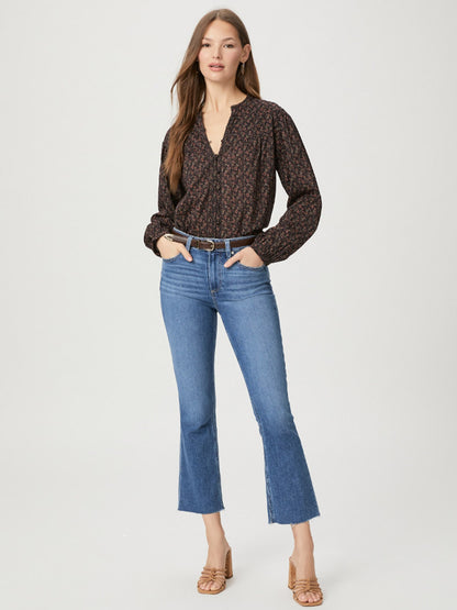 A woman stands wearing Paige Colette Crop in Starlet with a raw hem and a dark patterned blouse, with her hands slightly tucked into her pockets, against a plain background.