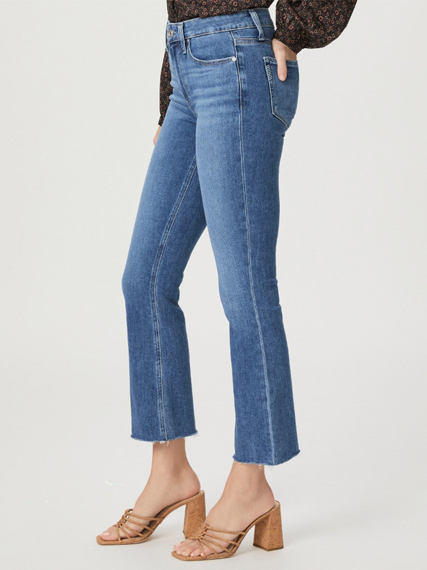 Woman standing sideways wearing blue Paige Colette Crop in Starlet denim high-rise cropped flare jeans with raw hem and brown heeled sandals, focusing on the outfit details from waist to feet.