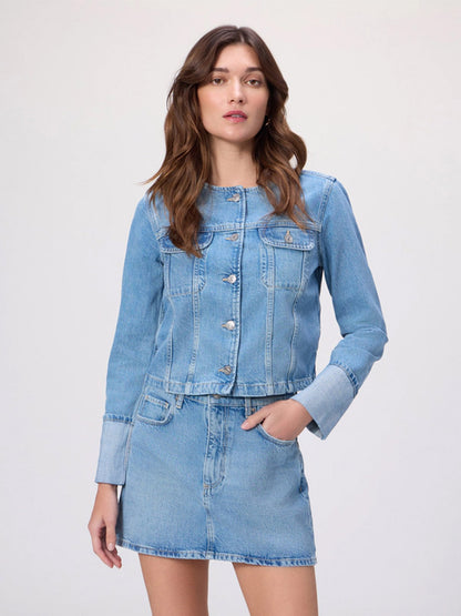 A woman stands against a plain background wearing the Paige Dean Jacket in Del Amo, a light wash collarless denim jacket, paired with a matching denim skirt and one hand in her skirt pocket.