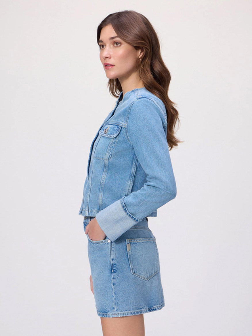 A woman stands in profile against a plain background, wearing the Paige Dean Jacket in Del Amo—a collarless, light wash denim jacket—and a matching skirt, with her left hand in her skirt pocket, exuding premium comfort.