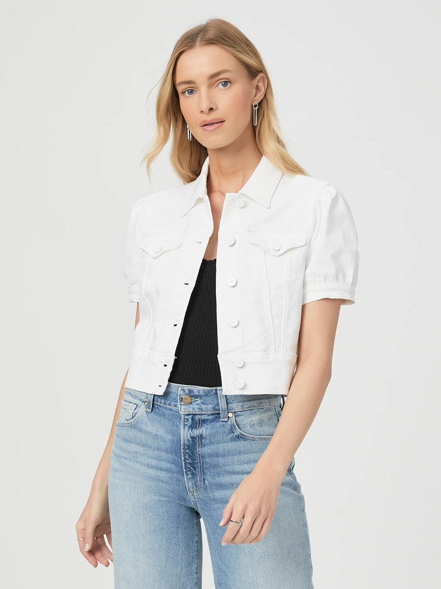 A woman wears the Paige Kendra Jacket in Crisp White, a cropped white denim jacket, over a black top and light blue jeans against a plain background.