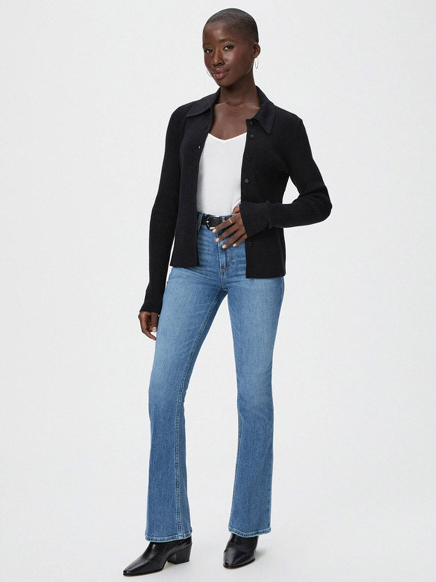 A person stands wearing a black cardigan, white top, and vintage denim blue jeans from the Paige Laurel Canyon High Rise Bootcut in Rock Show Distressed collection, paired with black boots against a plain white background.