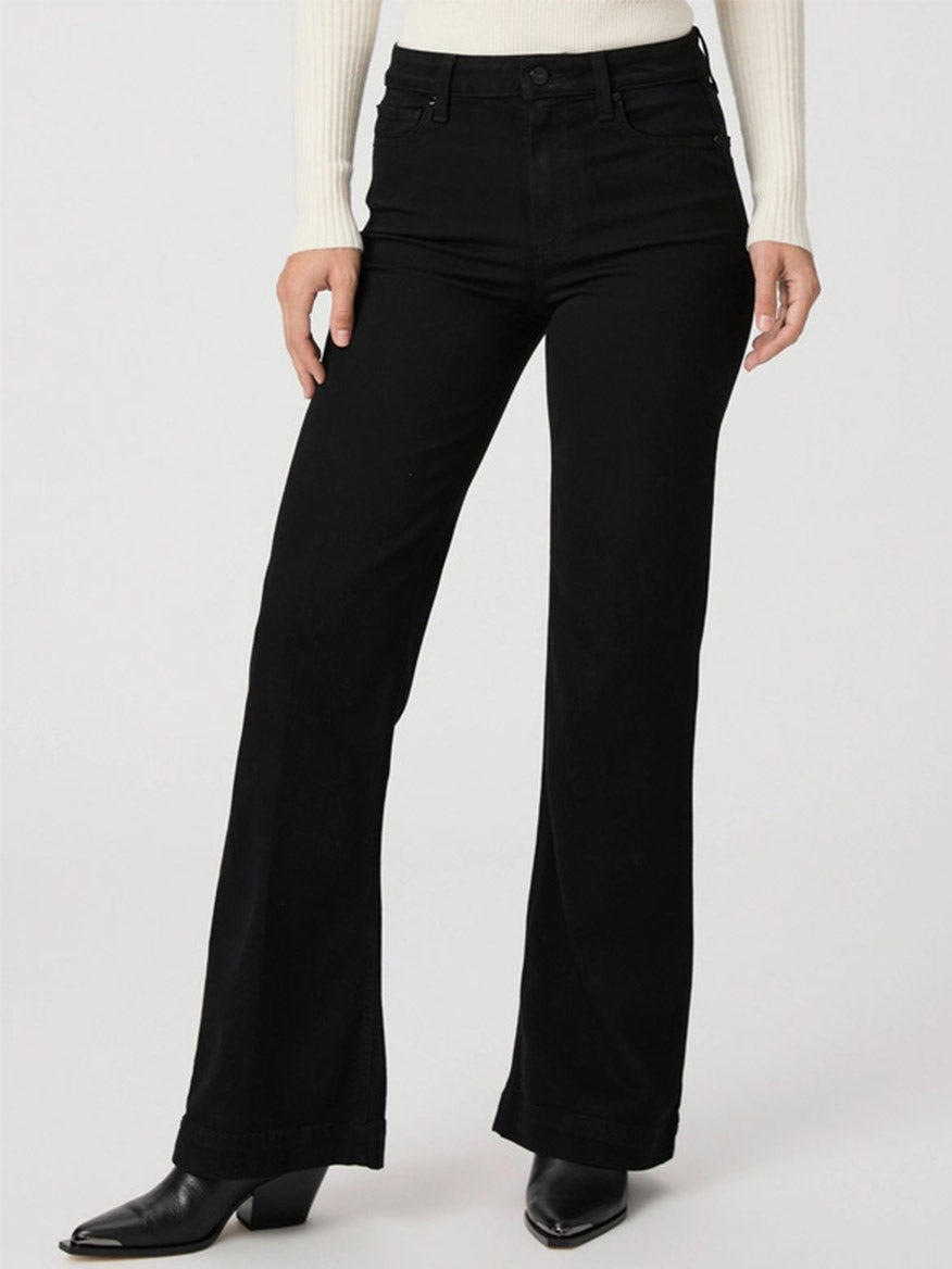 Person wearing a white long-sleeve sweater, Paige Leenah 32" Wide Leg Jeans in Black Shadow, and black heeled shoes stands against a plain background.