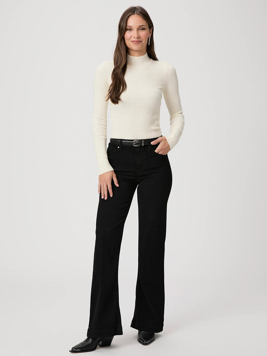 A person with long hair wearing a white ribbed turtleneck sweater, Paige Leenah 32" Wide Leg Jeans in Black Shadow, and black shoes is standing against a plain white background.