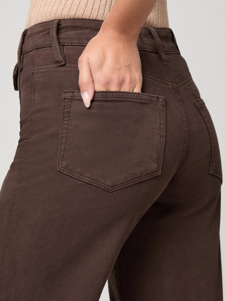 A person wearing the Paige Leenah 32″ Wide Leg Jean in Rich Chocolate with a beige ribbed top, their hand resting in the back pocket.