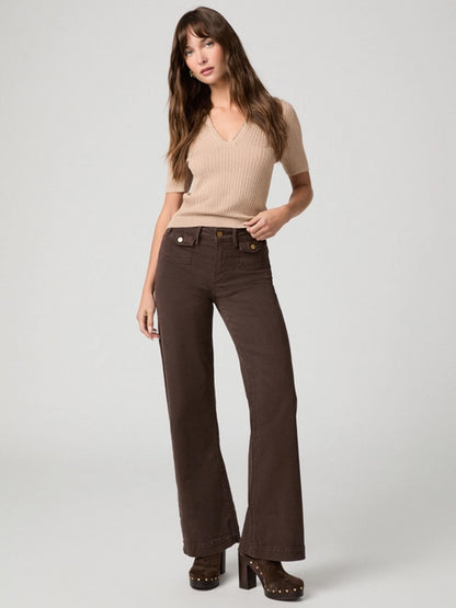 A woman stands against a plain background, wearing a beige short-sleeve sweater, Paige Leenah 32″ Wide Leg Jean in Rich Chocolate with utility patch pockets, and brown heeled clogs.