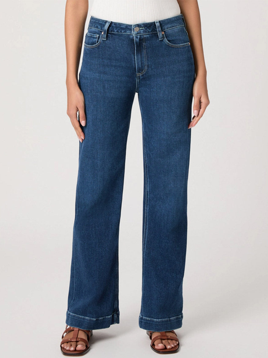 A person wearing Paige Leenah 32" Wide Leg Jean in Venetian Breeze, a white ribbed top, and brown sandals stands against a plain light background.