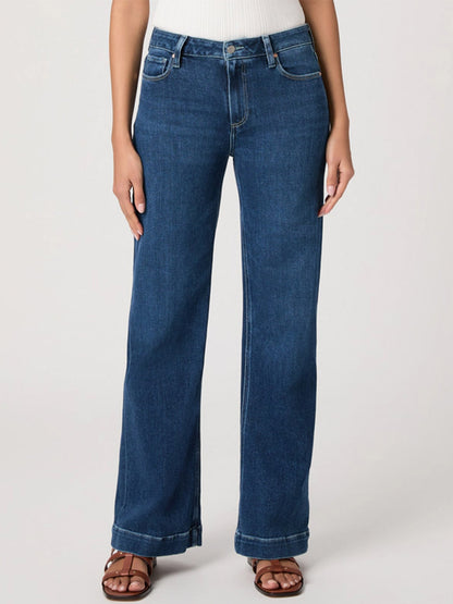 A person wearing Paige Leenah 32" Wide Leg Jean in Venetian Breeze, a white ribbed top, and brown sandals stands against a plain light background.