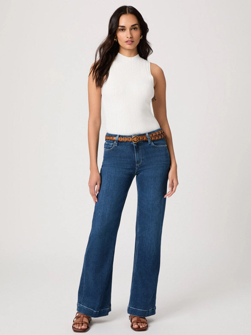 Woman standing against a plain background, wearing a sleeveless white top, Paige Leenah 32" Wide Leg Jean in Venetian Breeze with a brown belt, and brown sandals.