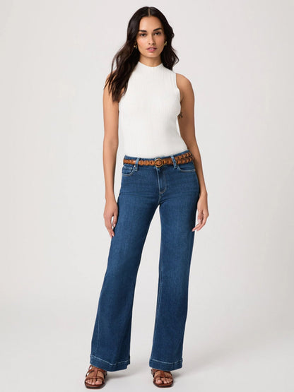 Woman standing against a plain background, wearing a sleeveless white top, Paige Leenah 32" Wide Leg Jean in Venetian Breeze with a brown belt, and brown sandals.