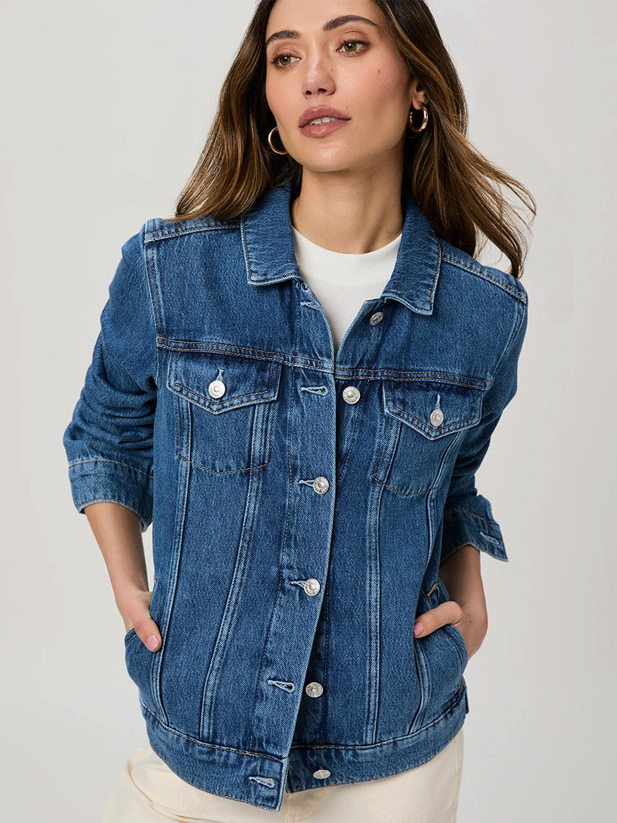 Woman wears the Paige Rowan Jacket in Bellisimo, a medium wash denim piece, over a white top with hands in pockets, standing against a light background for a fresh, vintage vibe.
