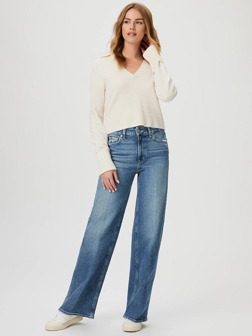 A person wearing a white sweater and blue Paige Sasha 32" Wide Leg Jean in Storybook Distressed stands against a plain background.