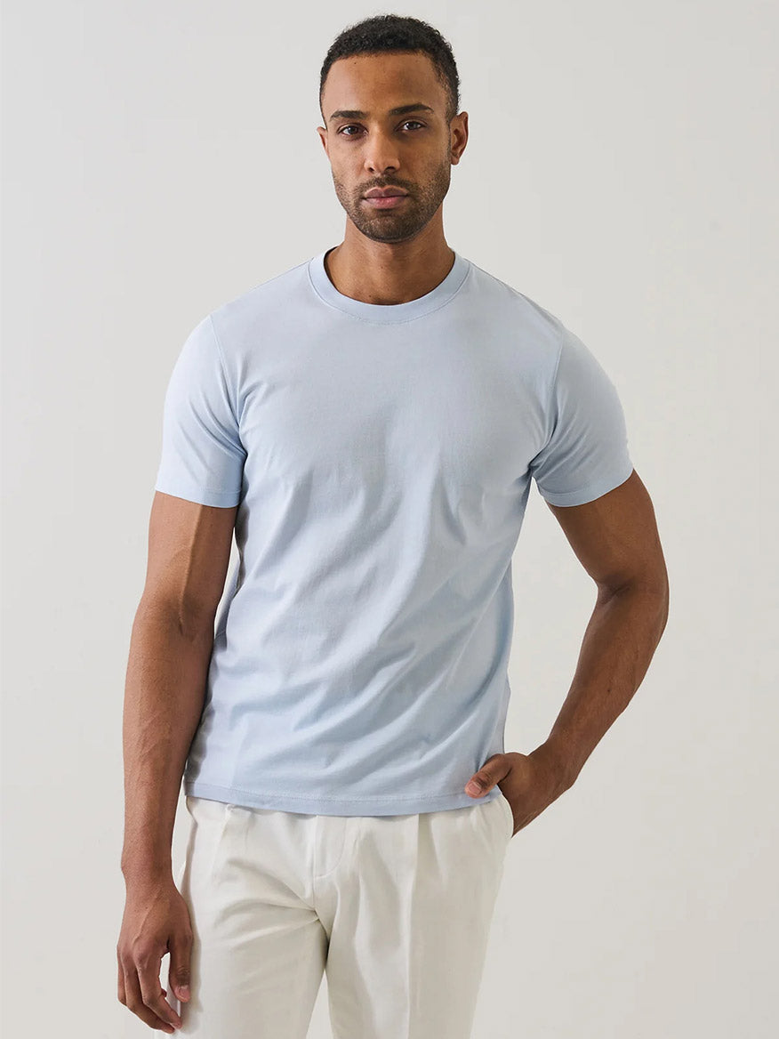 A man stands against a plain background wearing the Patrick Assaraf Organic Pima Cotton High Neck T-Shirt in Blue Ice and white pants, with one hand in his pocket.