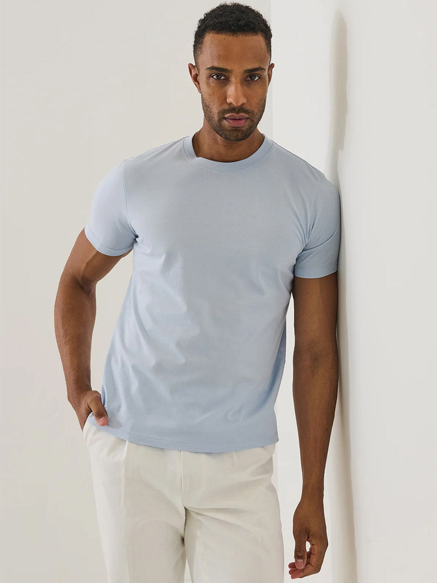 A man stands against a white wall, wearing the Patrick Assaraf Organic Pima Cotton High Neck T-Shirt in Blue Ice and white pants, with one hand in his pocket.