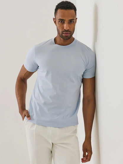 A man stands against a white wall, wearing the Patrick Assaraf Organic Pima Cotton High Neck T-Shirt in Blue Ice and white pants, with one hand in his pocket.
