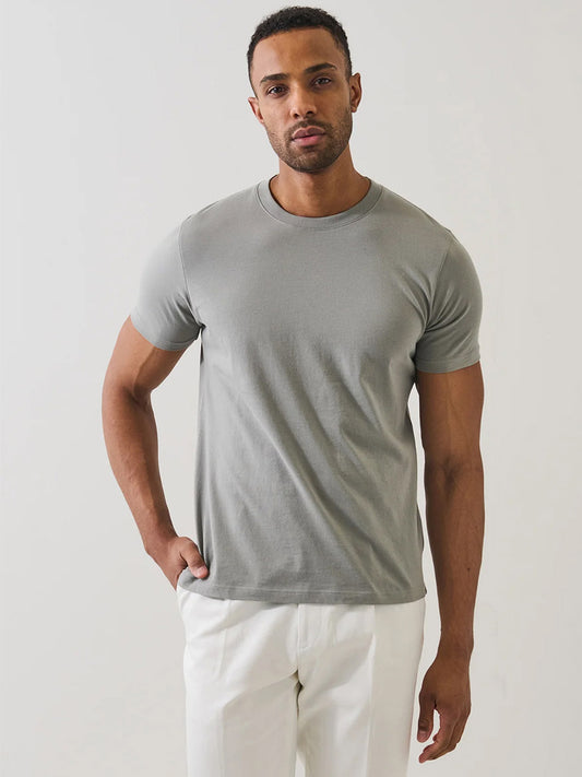 A man stands against a plain background, wearing the Patrick Assaraf Organic Pima Cotton High Neck T-Shirt in Pewter and white pants—an effortlessly stylish statement of sustainable fashion.
