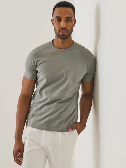 A man in a pewter Patrick Assaraf Organic Pima Cotton High Neck T-Shirt and white pants stands against a white wall, looking at the camera with one hand in his pocket—a symbol of effortless, sustainable style.