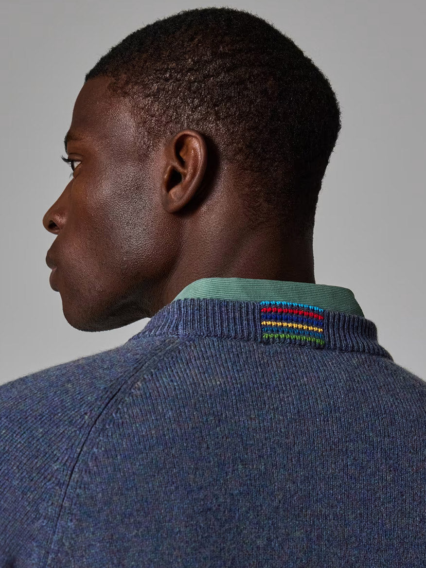 A man is shown from the back and side, wearing a Paul Smith Indigo Merino Wool Crew Neck Sweater with a green shirt collar and a small multicolored embroidered patch near the neckline.