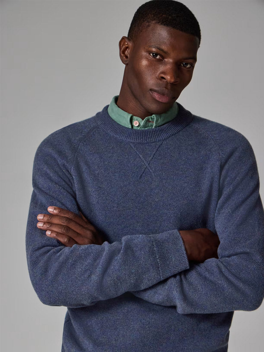 A man stands with arms crossed, wearing a Paul Smith Indigo Merino Wool Crew Neck Sweater over a green button-up shirt, looking directly at the camera against a plain gray background.