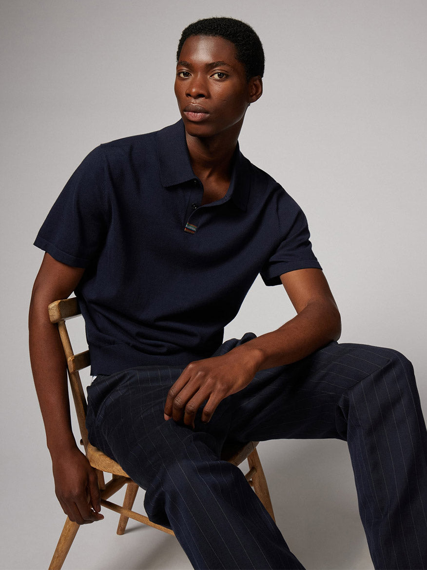 Seated on a wooden chair, the person wears the Paul Smith Knitted Cotton 'Signature Stripe' Trim Polo Shirt in Navy with dark pinstriped trousers, gazing at the camera before a plain backdrop; a subtle Signature Stripe elevates the look.