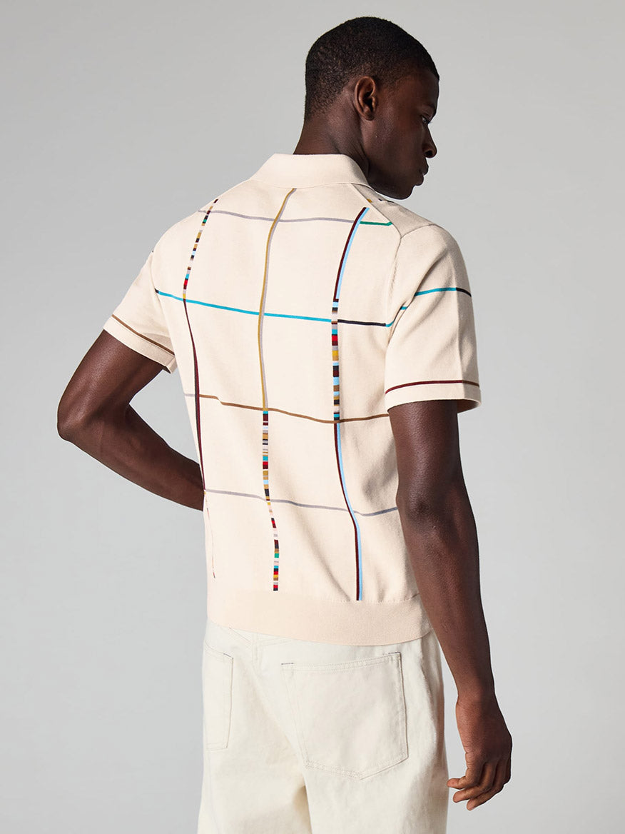 A man wearing a Paul Smith Signature Stripe Check Knitted Cotton Polo Shirt in Ecru, featuring a multicolored grid pattern on the back, pairs it with light pants and stands facing away from the camera.