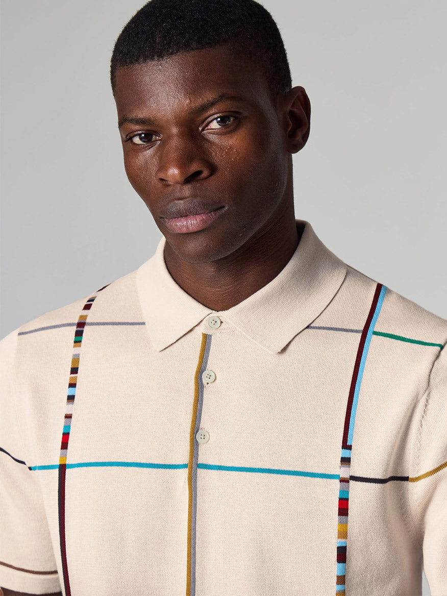 A man stands against a grey background wearing the Paul Smith Signature Stripe Check Knitted Cotton Polo Shirt in Ecru.