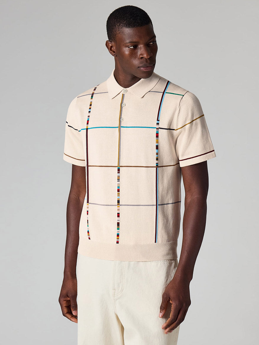 A man wearing a Paul Smith Signature Stripe Check Knitted Cotton Polo Shirt in ecru and cream-colored pants stands against a plain light gray background.