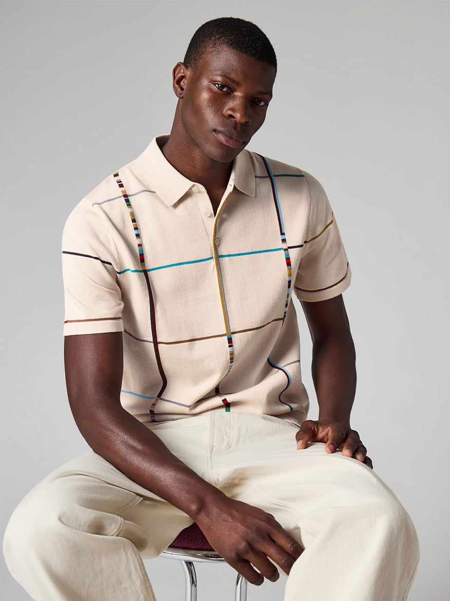 Man sitting on a stool wearing the Paul Smith Signature Stripe Check Knitted Cotton Polo Shirt in Ecru, paired with light-colored pants against a plain background.
