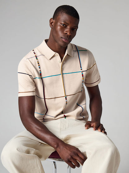 Man sitting on a stool wearing the Paul Smith Signature Stripe Check Knitted Cotton Polo Shirt in Ecru, paired with light-colored pants against a plain background.