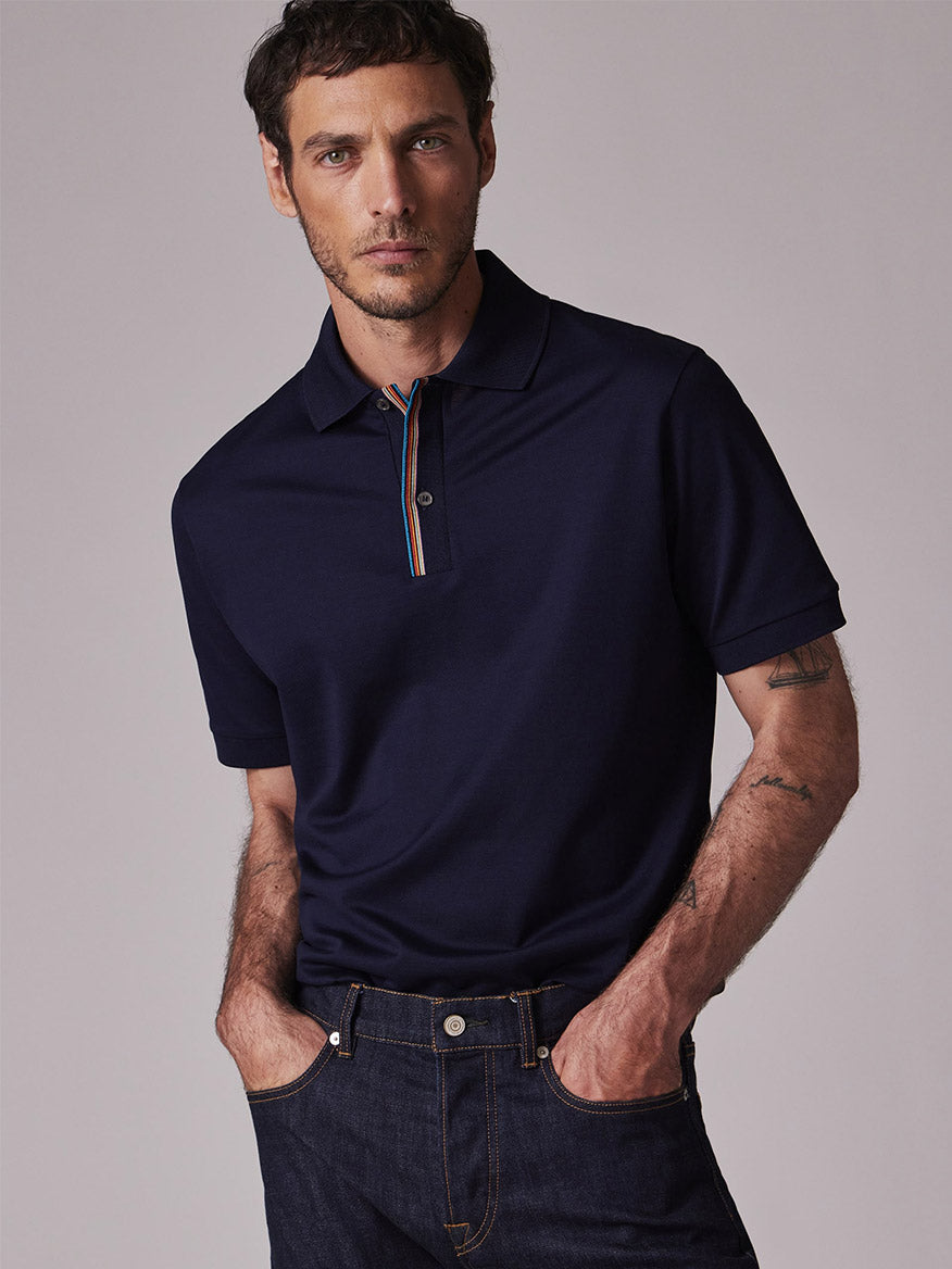 Man wearing a Paul Smith Signature Stripe Trim Polo Shirt in Dark Navy, featuring rainbow trim on the placket and paired with dark blue jeans, standing against a plain background.