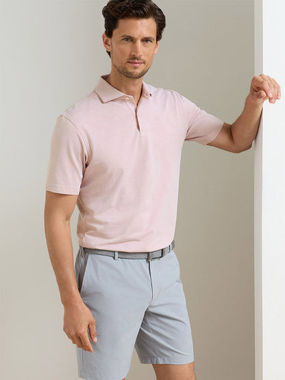 Man wearing a Peter Millar Albatross Cotton Blend Piqué Polo in Faded Scarlet, tucked into light gray shorts, standing indoors and leaning against a white wall.