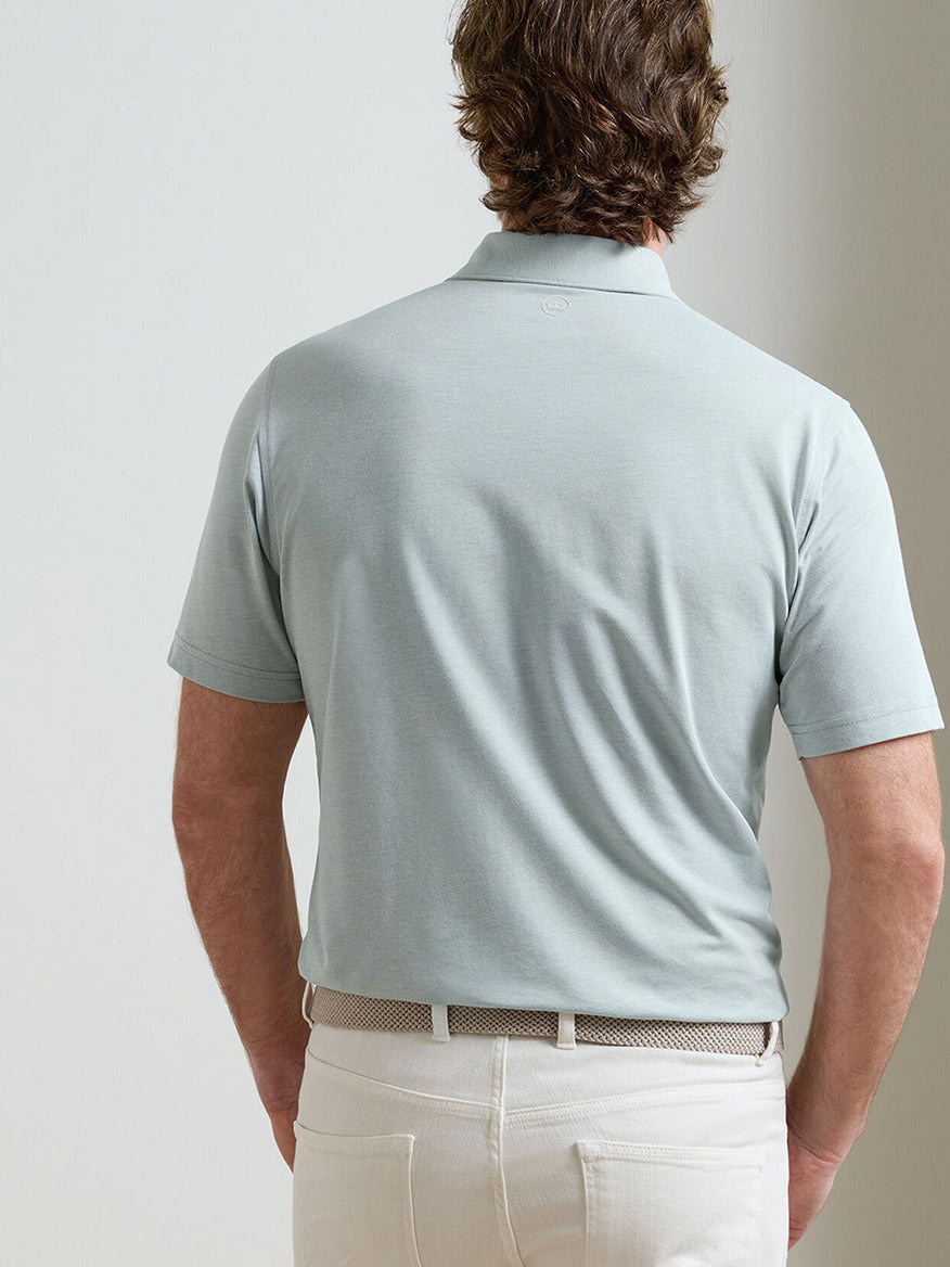A man stands facing away from the camera, wearing a Peter Millar Albatross Cotton Blend Piqué Polo in Light Willow and off-white pants with a textured belt.