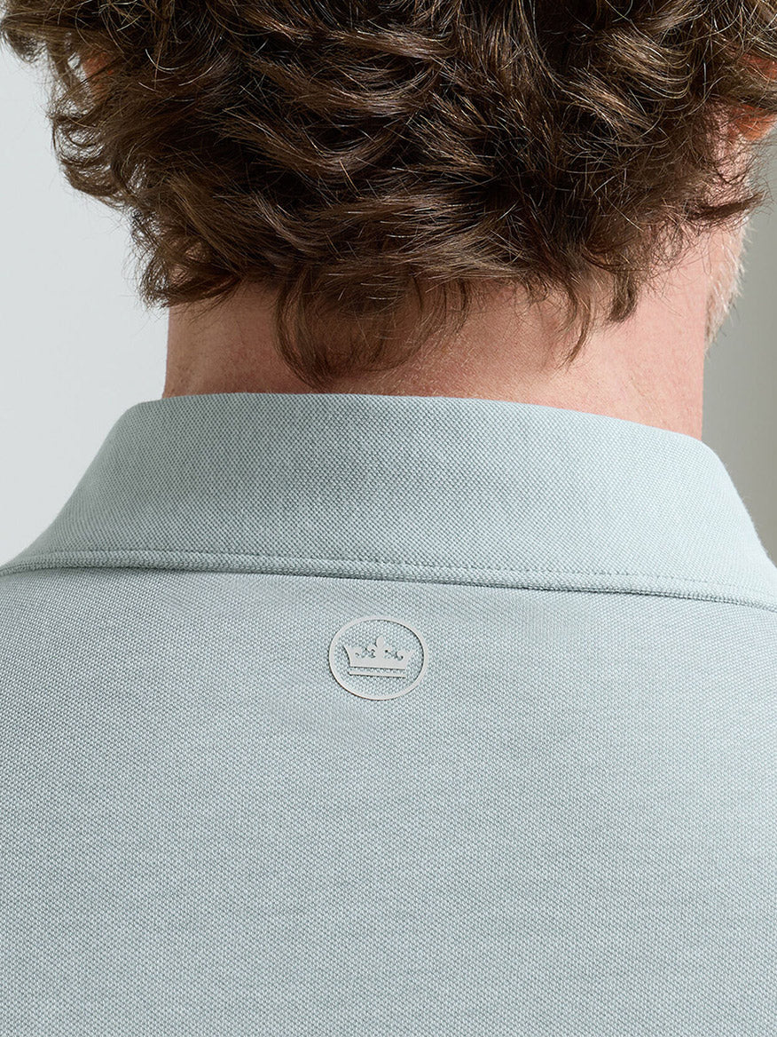 Close-up of the back of a person's neck and shoulders, showcasing the Peter Millar Albatross Cotton Blend Piqué Polo in Light Willow, with a light gray hue and small circular logo beneath the collar.