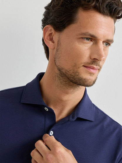 A man with short, wavy brown hair looks to the side while buttoning the collar of a Peter Millar Albatross Cotton Blend Piqué Polo in Navy.
