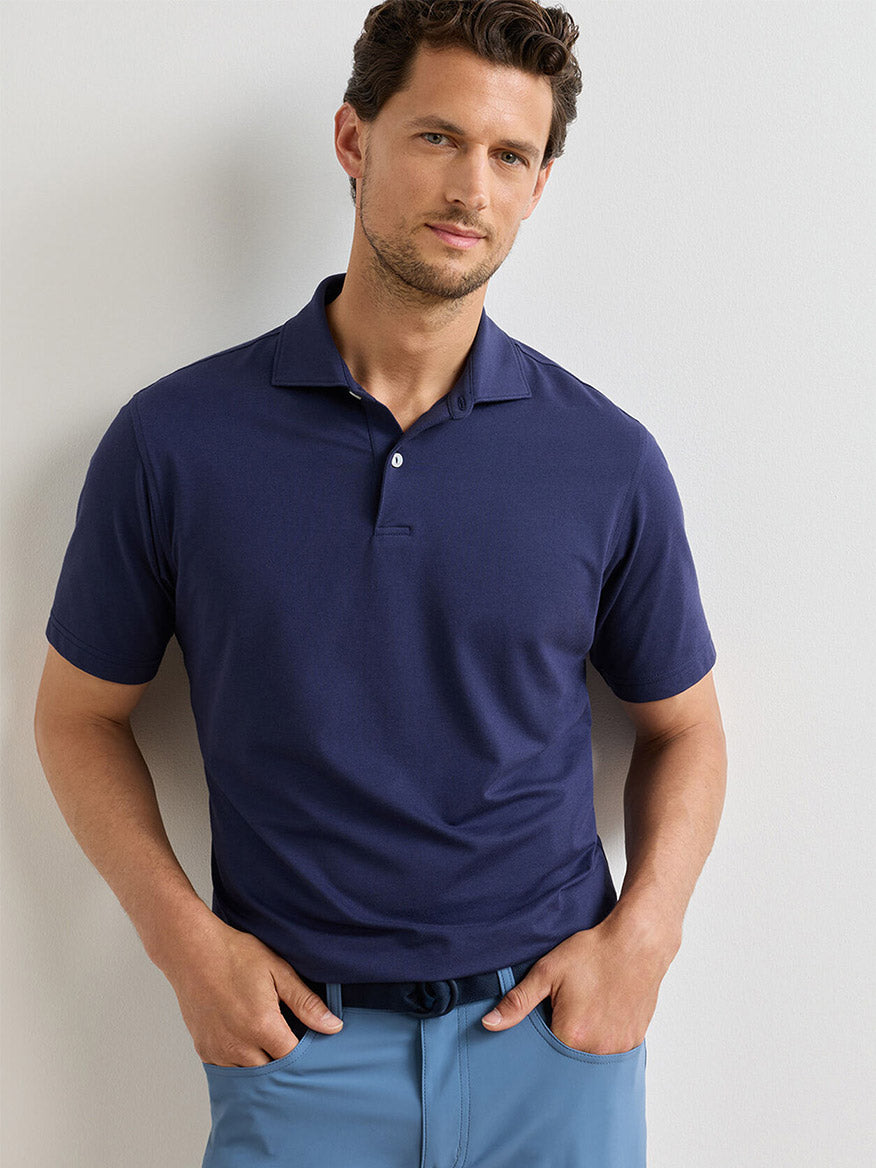 A man wearing the Peter Millar Albatross Cotton Blend Piqué Polo in Navy and light blue pants stands against a plain light-colored wall, facing the camera with his hands in his pockets.