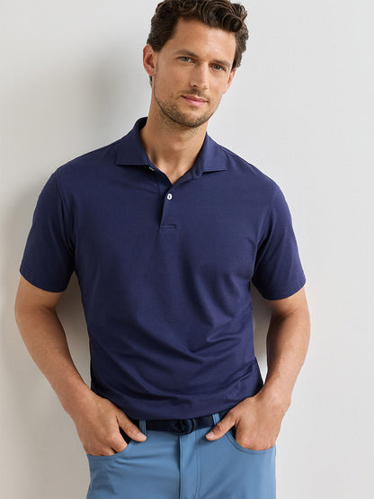 A man wearing the Peter Millar Albatross Cotton Blend Piqué Polo in Navy and light blue pants stands against a plain light-colored wall, facing the camera with his hands in his pockets.