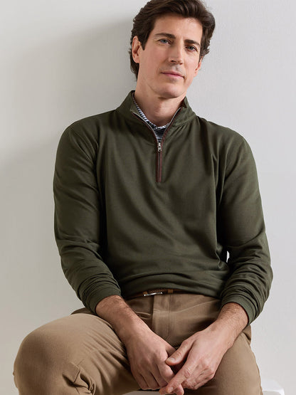 A man with short brown hair is wearing the Peter Millar Albatross Cotton-Blend Piqué Quarter-Zip in Dark Olive, designed for performance, paired with tan pants as he sits against a white wall with his arms resting on his legs.