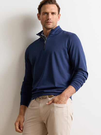 A man wearing the Peter Millar Albatross Cotton-Blend Piqué Quarter-Zip in Navy and beige pants stands against a plain white wall, one hand in his pocket.