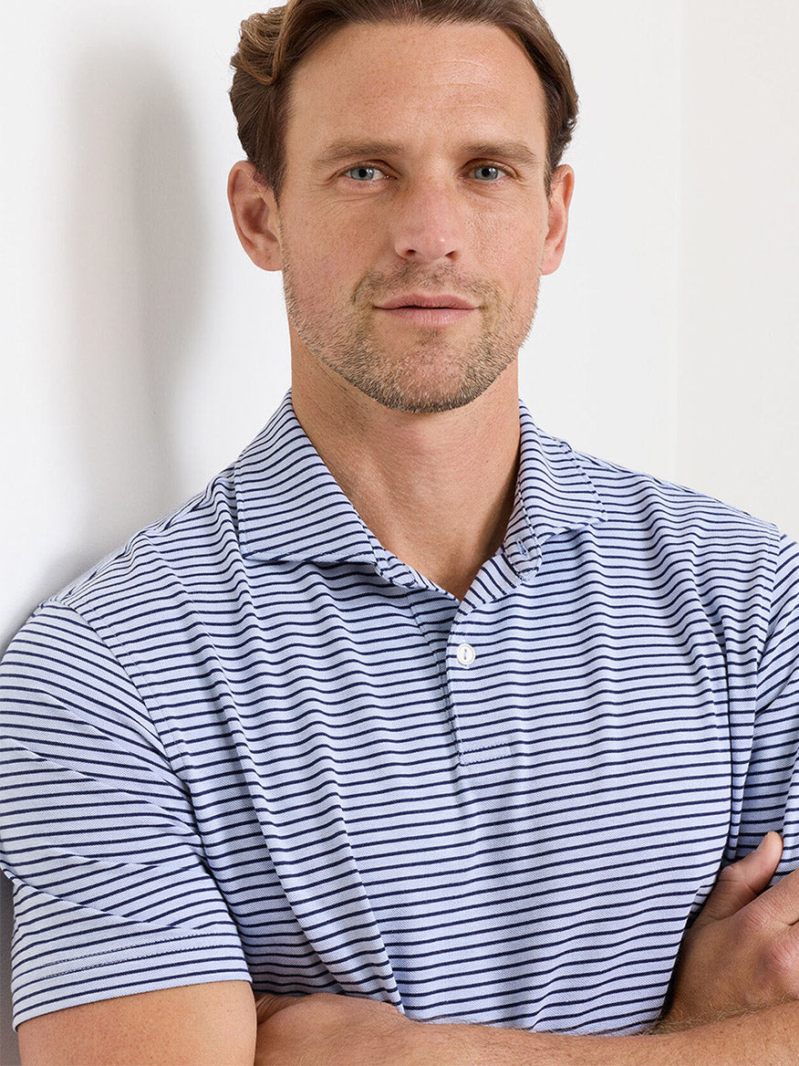 Man with short brown hair and light stubble wears the Peter Millar Albatross Stripe Cotton-Blend Piqué Polo in Dusk Blue, short-sleeved with white and blue stripes, standing with arms crossed and looking at the camera.