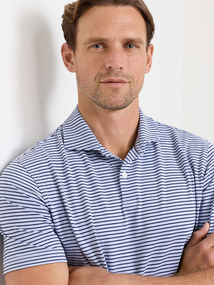 Man with short brown hair and light stubble wears the Peter Millar Albatross Stripe Cotton-Blend Piqué Polo in Dusk Blue, short-sleeved with white and blue stripes, standing with arms crossed and looking at the camera.
