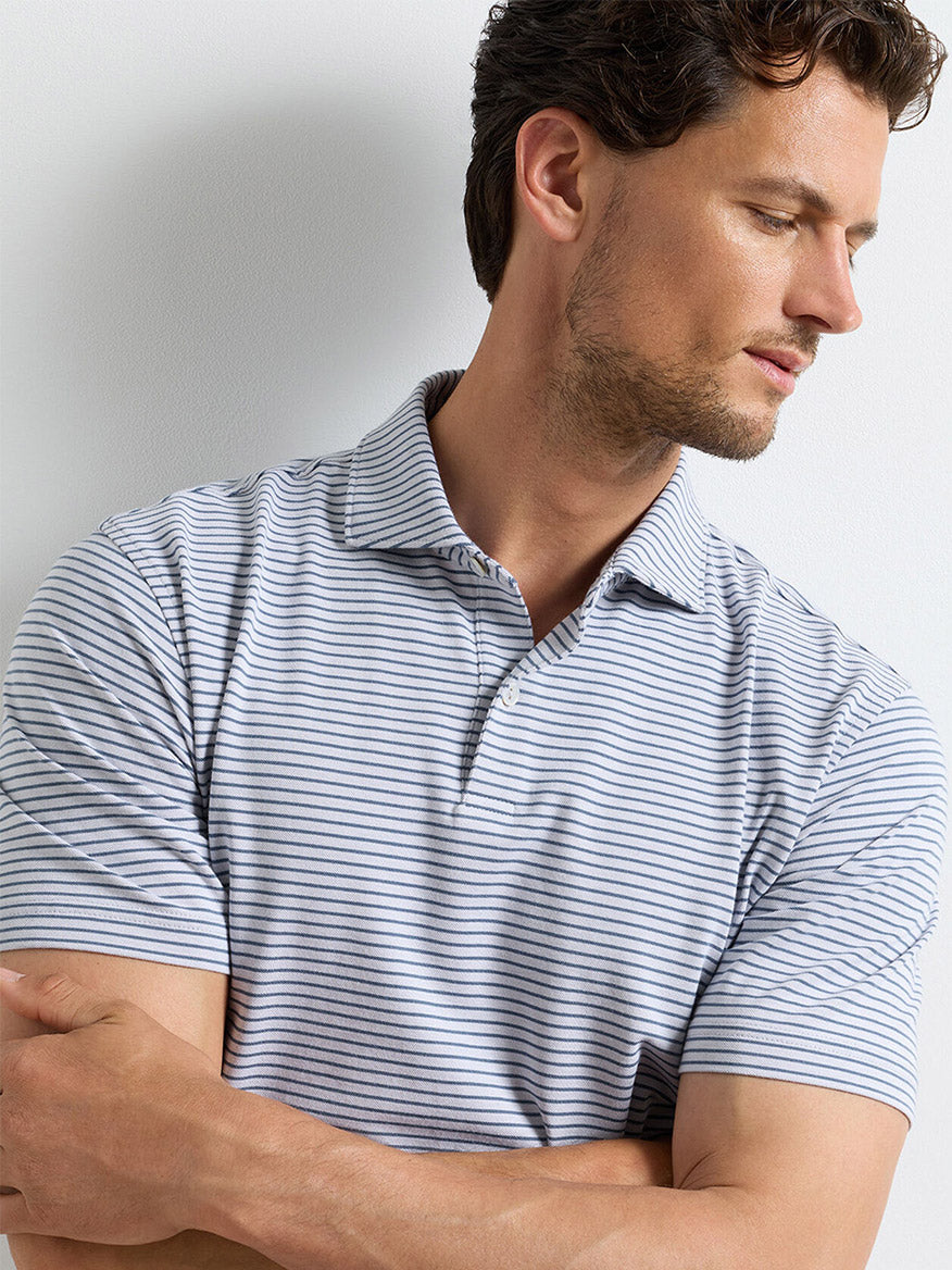 Man wearing a Peter Millar Albatross Stripe Cotton-Blend Piqué Polo in Gale Grey with short sleeves and thin black stripes, arms loosely crossed, looking down to his left.