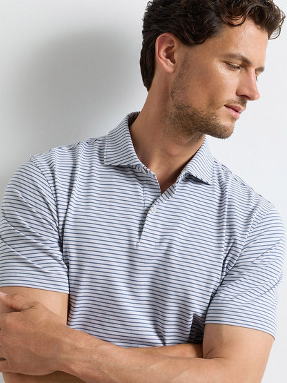 Man wearing a Peter Millar Albatross Stripe Cotton-Blend Piqué Polo in Gale Grey with short sleeves and thin black stripes, arms loosely crossed, looking down to his left.