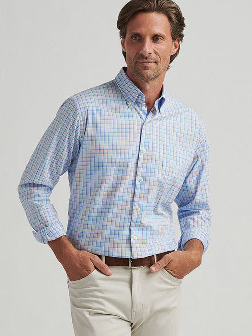 A man wearing a Peter Millar Albert Crown Lite Cotton-Stretch Sport Shirt in Blue Poppy, featuring a light blue checkered pattern and rolled-up sleeves, stands with hands in pockets against a plain background, highlighting the classic fit and easy-care fabric.