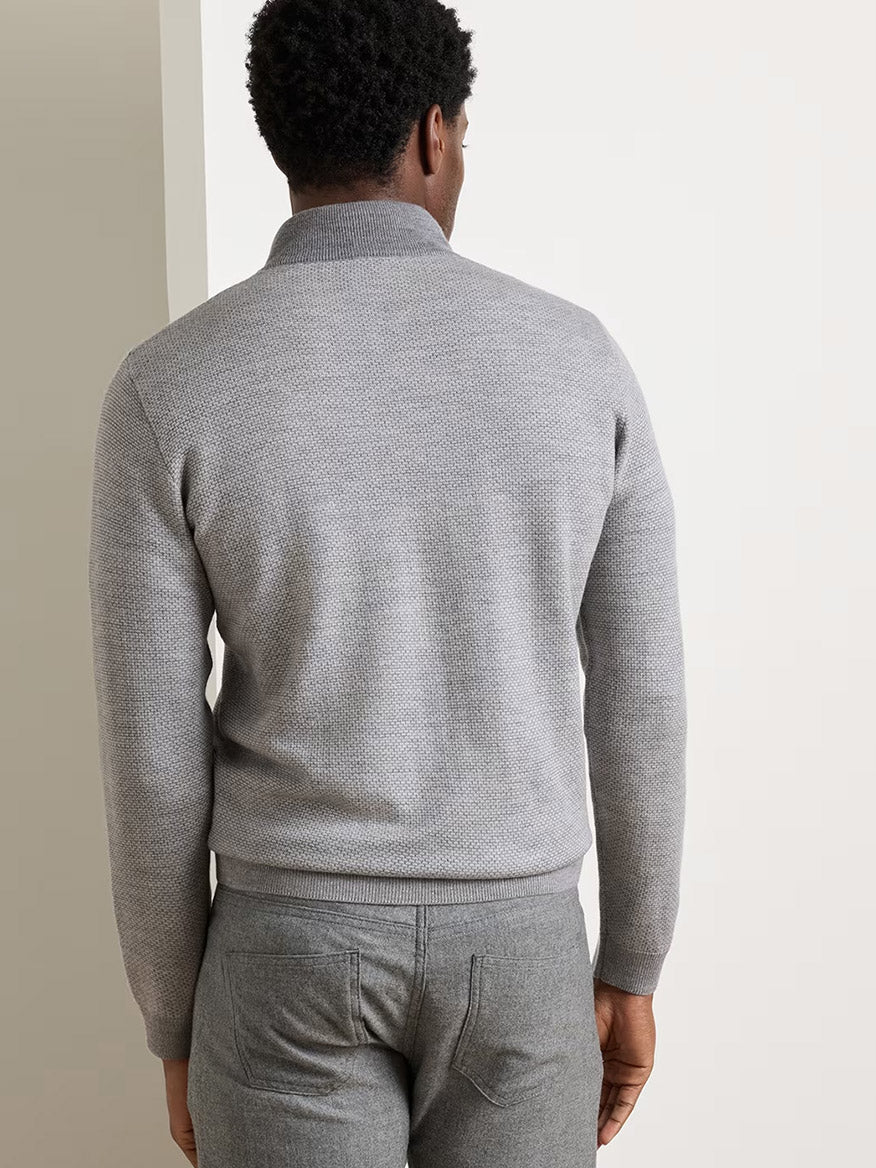 A person stands facing away from the camera, wearing a Peter Millar Ashland Quarter-Zip Sweater in Gale Grey paired with matching gray pants, in front of a neutral background.