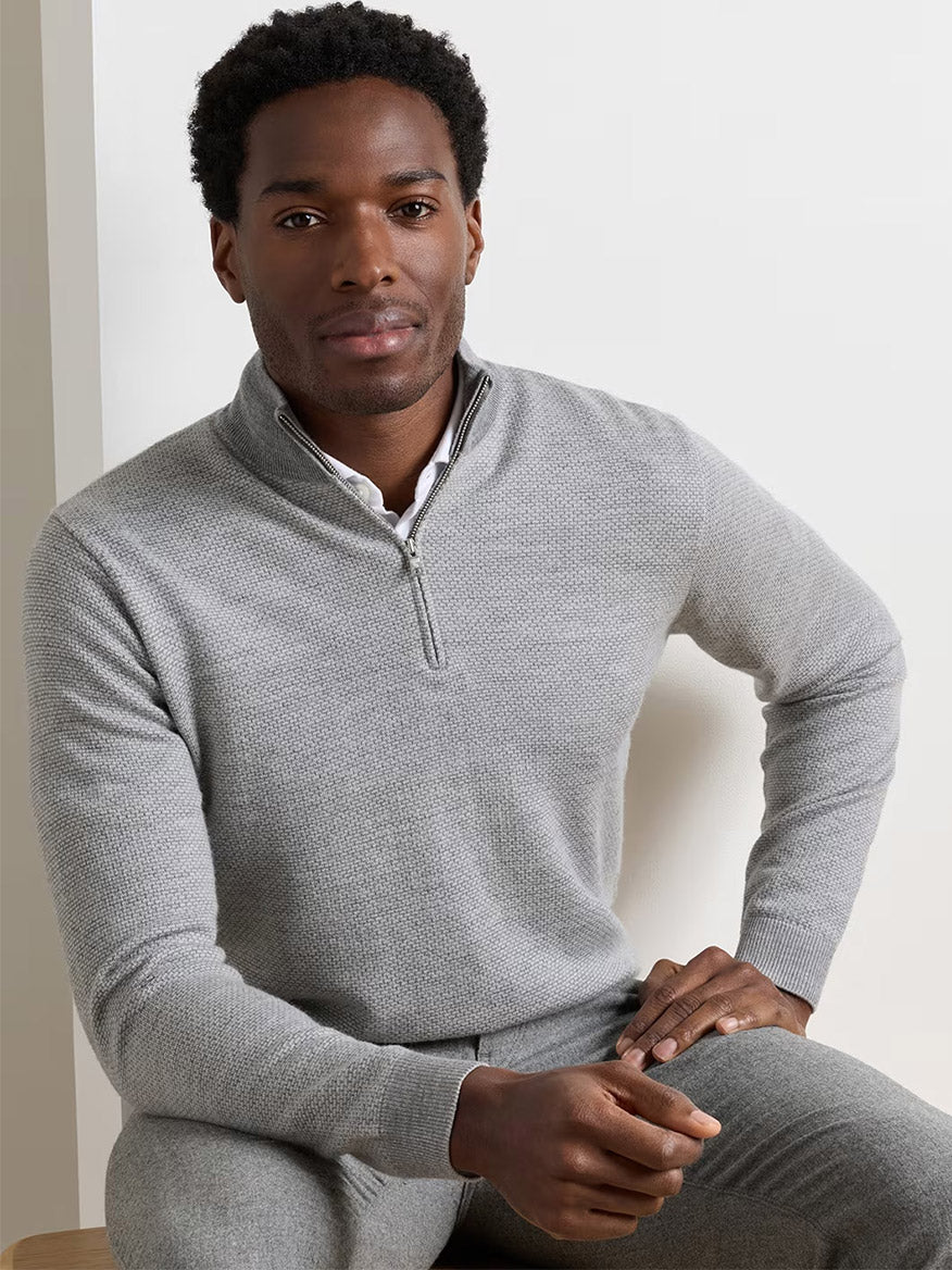 A man sits on a stool against a plain background, wearing the Peter Millar Ashland Quarter-Zip Sweater in Gale Grey—a tailored-fit, light gray midweight sweater with a subtle jacquard pattern—layered over a white shirt and paired with matching gray pants.