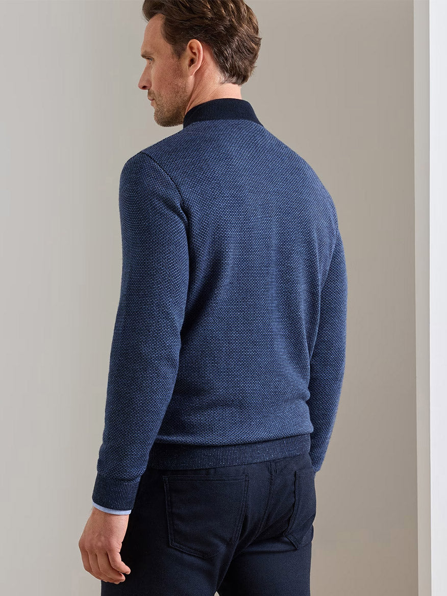 A man stands facing away, wearing the Peter Millar Ashland Quarter-Zip Sweater in Midnight Navy—a tailored fit sweater with a textured jacquard pattern, dark collar and cuffs—paired with black jeans, in a neutral indoor setting.